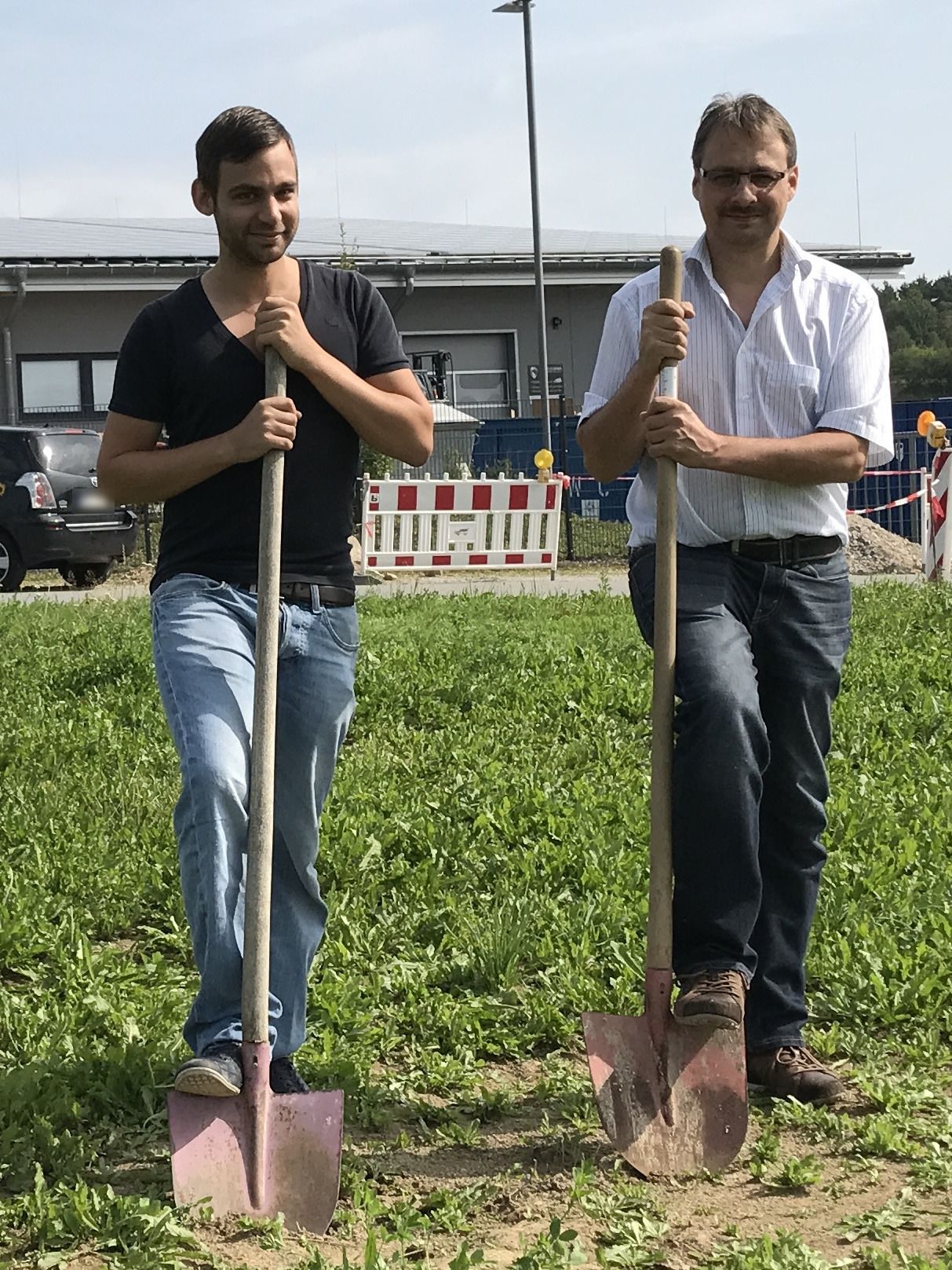 Zwei Männer stehen mit Spaten auf einer Wiese, im Hintergrund eine Baustellenabsperrung und ein Gewerbegebäude.